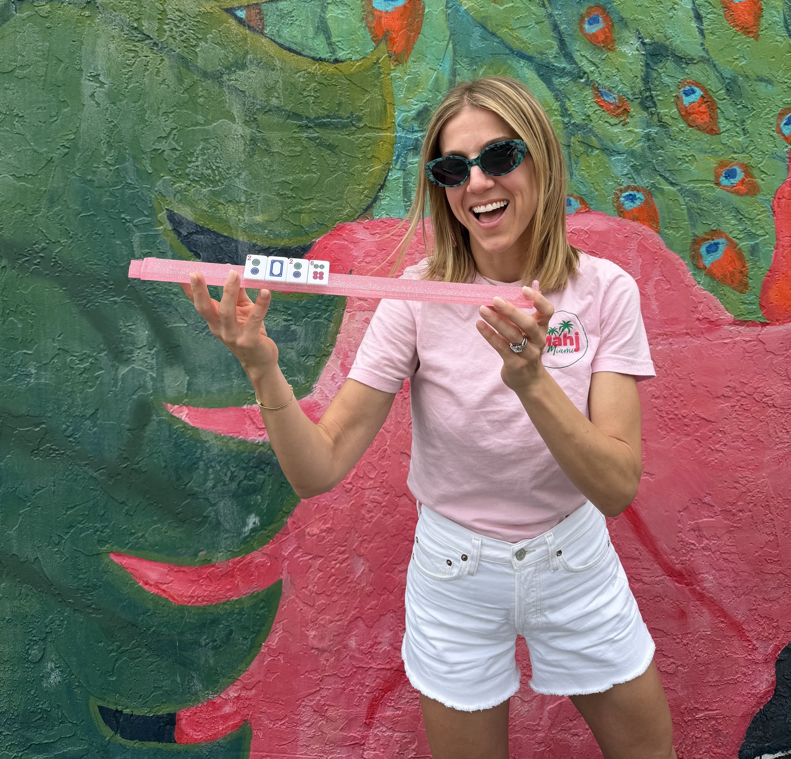 Abby laughing and holding a pink mahjong tile rack in front of a colorful Miami peacock mural